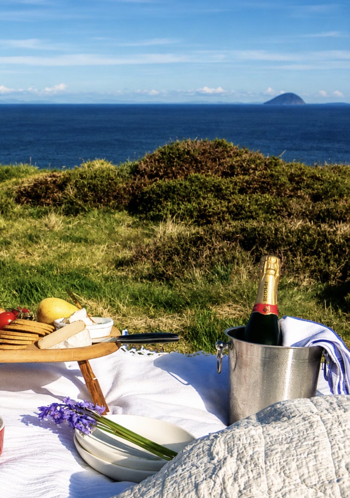 An island picnic with champagne laid out on a rug above the sea